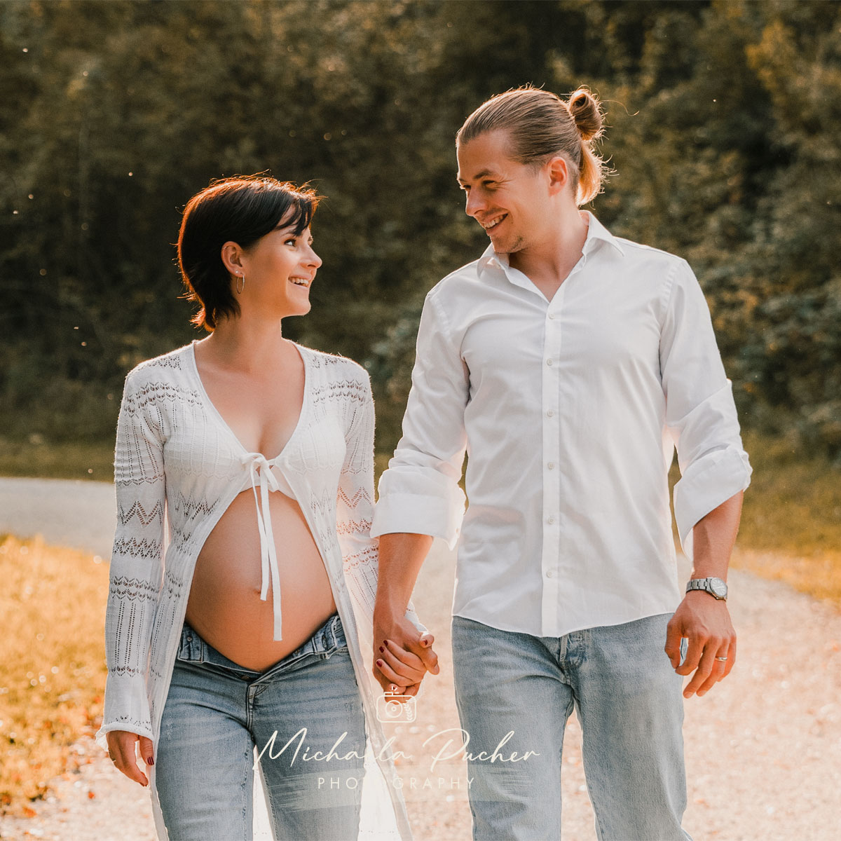 Schwangere Frau geht Hand in Hand mit ihrem Partner auf einem sonnigen Weg, beide lächeln sich beim Gehen fröhlich an; sie trägt Jeans und eine offene Strickjacke, er ein weißes Hemd.